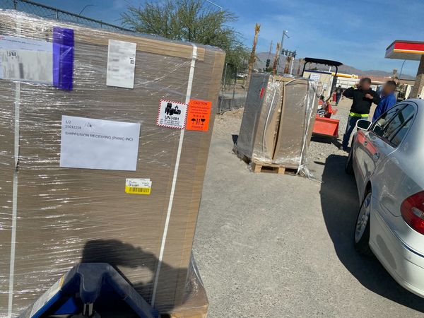 Two pallets of Playdates addressed to Ship Fusion in an under-construction gas station parking lot. Two people stand nearby; their faces have been blurred out.